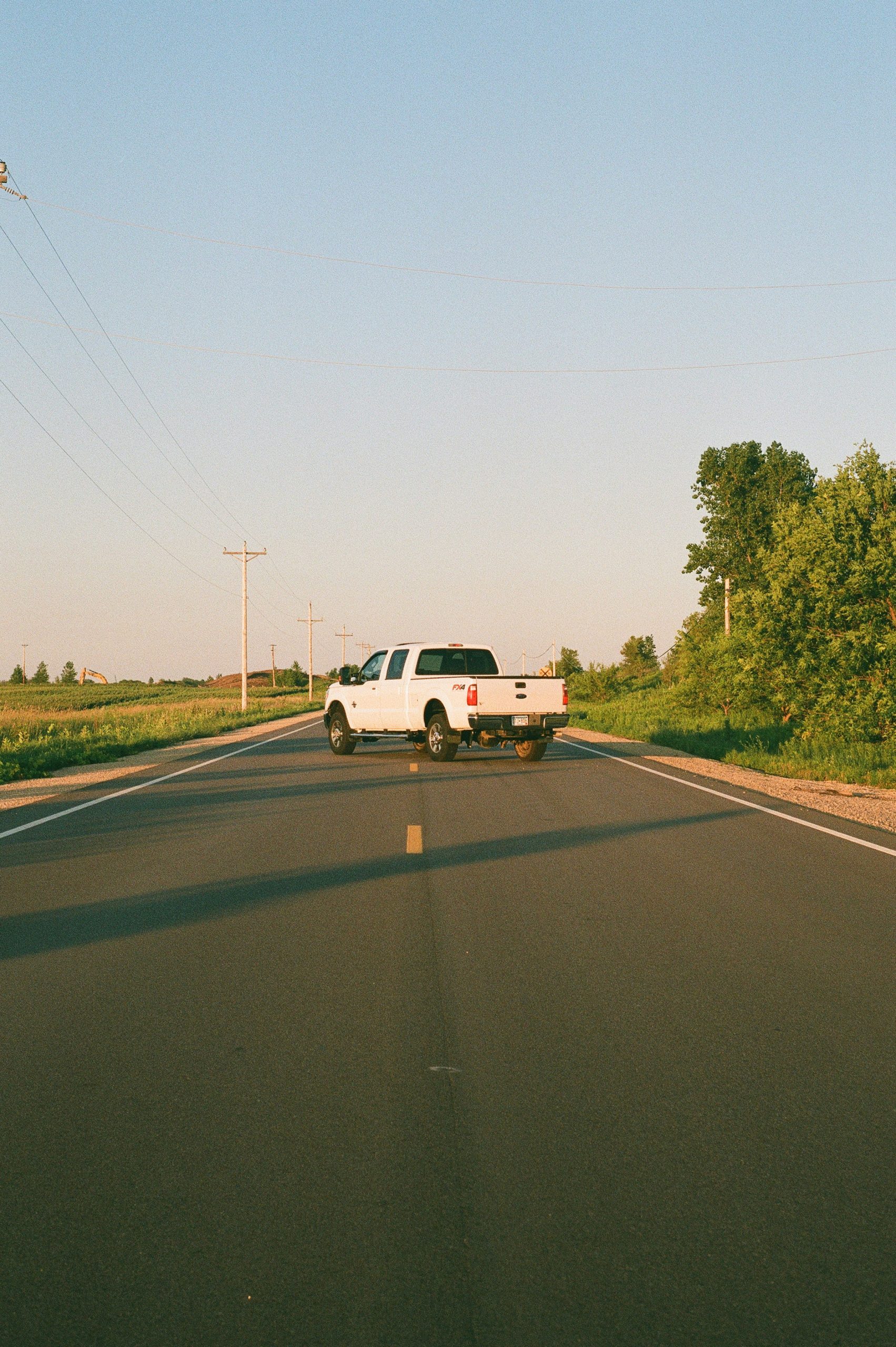 White pickup truck traveling a rural road lined with trees and power lines, capturing the scenic approach to a Kansas guided hunting lodge.